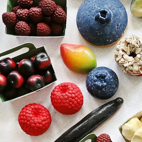 A colorful arrangement of realistic-looking fruits and desserts, including raspberries, blueberries, a mango, and a donut, on a patterned surface. At Le Meurice, Paris.