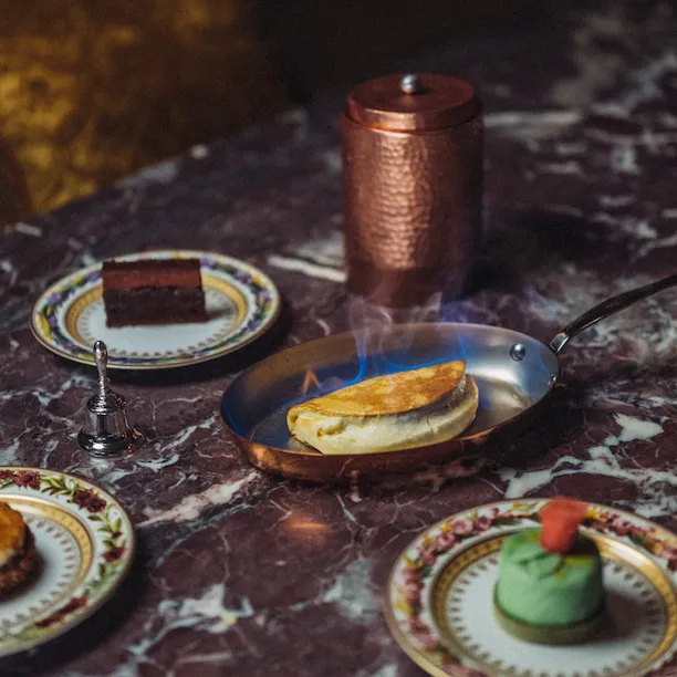 Dessert assortment on a marble table with a crêpe soufflée being flamed, chocolat charlotte, and Ambassadeur cake, at the Jean Imbert au Plaza Athénée, at Hotel Plaza Athénée, Paris