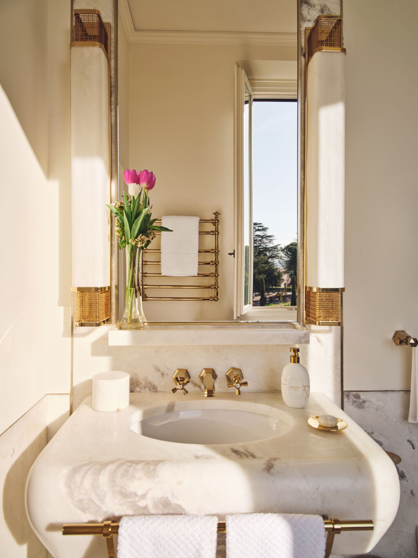 Detail of sink area with mirror reflecting the view from an open window in  a guest room bathroom at Hotel Eden Rome