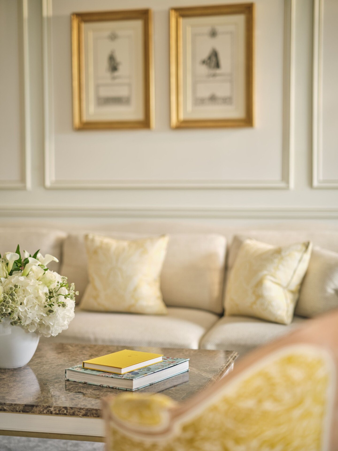Detail picture of a prestige suite. A marble table with a flower arrangement and two books in visible on the foreground, a sofa in soft beige colours is in the background, at Le Meurice, Paris. 