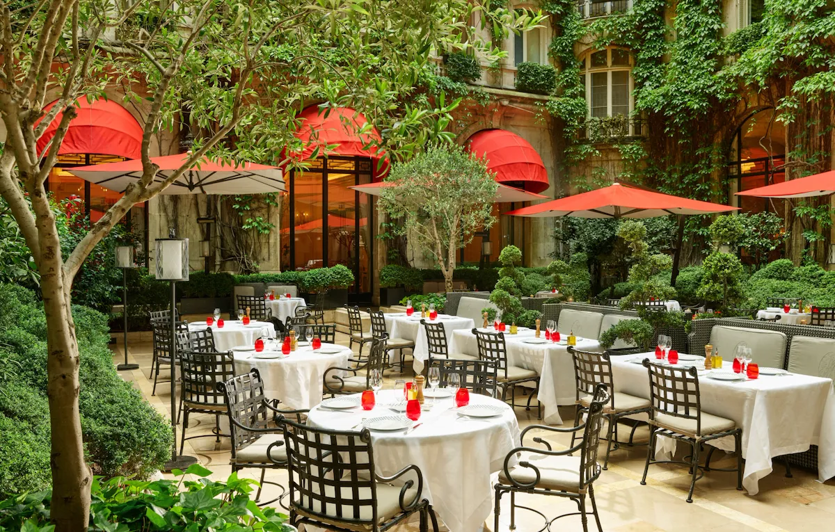 Terrace of La Cour Jardin with drawn up tables, iconic red parasols, green vine and trees seen from the side, at Hotel Plaza Athénée, Paris