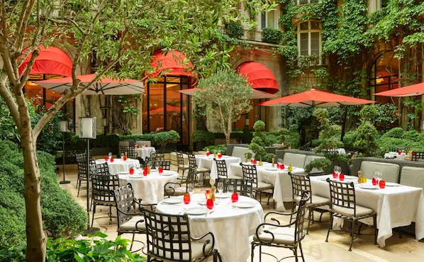 Terrace of La Cour Jardin with drawn up tables, iconic red parasols, green vine and trees seen from the side, at Hotel Plaza Athénée, Paris