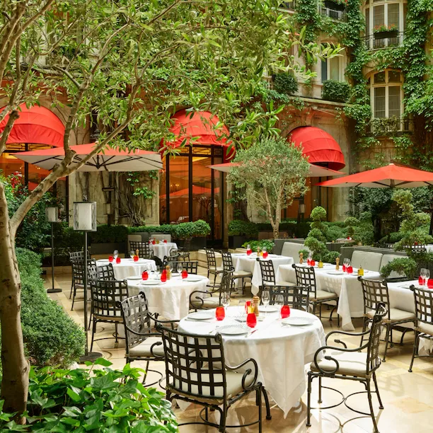 Terrace of La Cour Jardin with drawn up tables, iconic red parasols, green vine and trees seen from the side, at Hotel Plaza Athénée, Paris