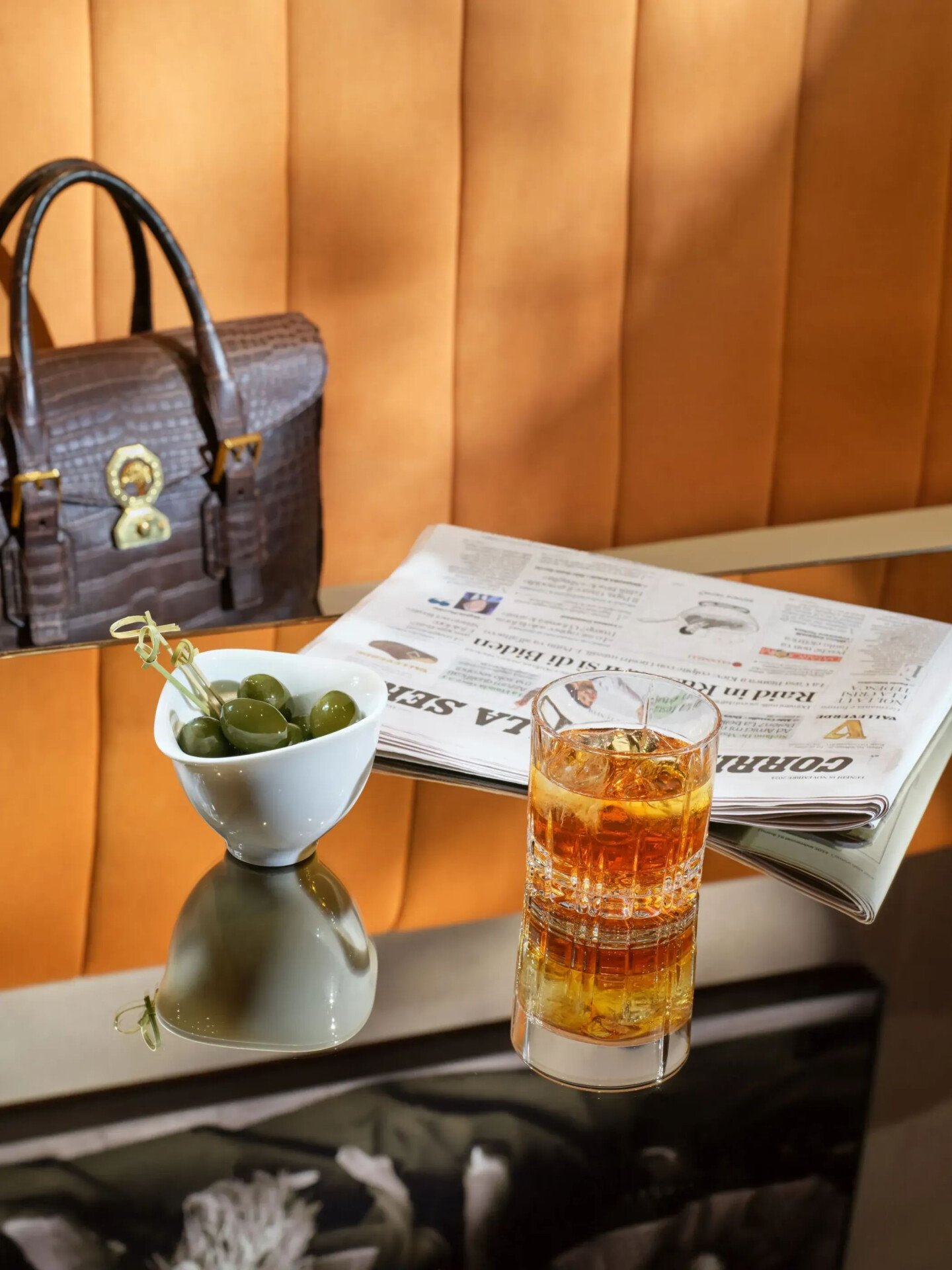 Close up on one of the tables in Il Salotto Lobby Lounge with a cocktail, green olives and a newspaper. Leather work bag in the background 