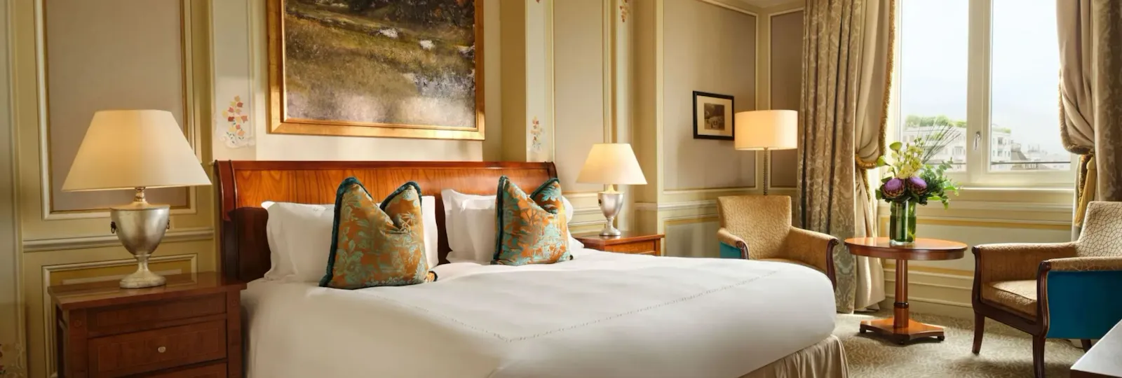 The room is decorated in soft gold colours and features decorative gold and blue cushions. There is a large painting above the bed and a large window that lets in plenty of natural light.