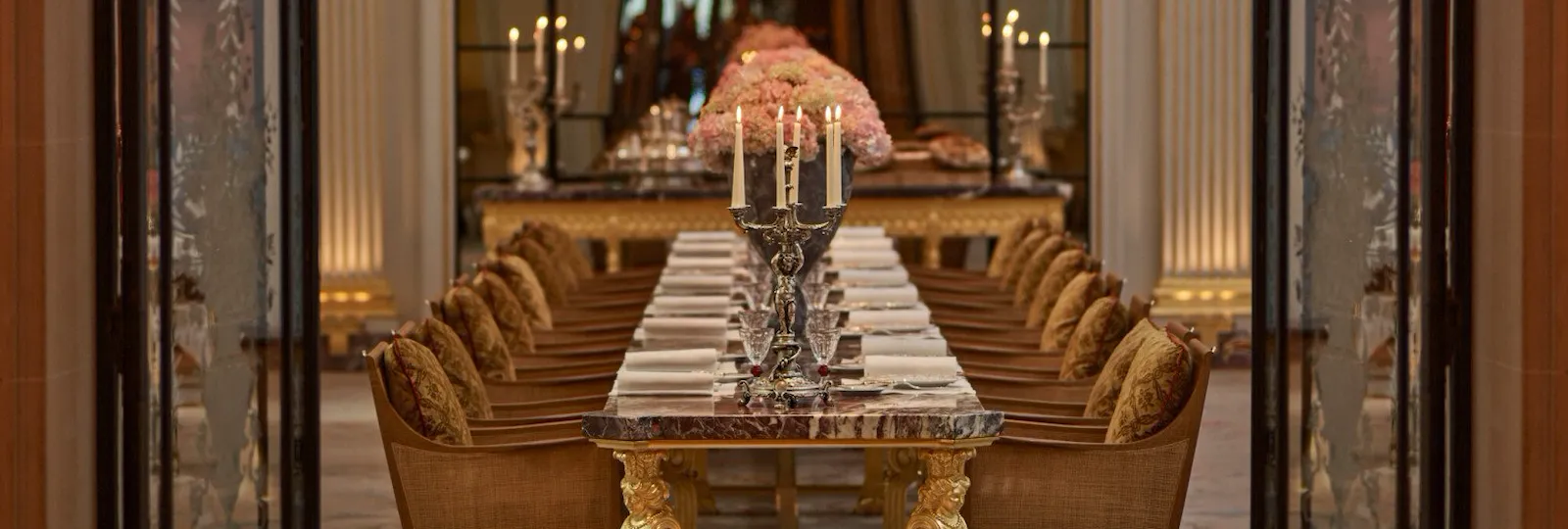 Jean Imbert au Plaza Athénée's central marble table, drawned up with pink flowers and chandeliers. Crystal chandeliers hanging above the table. Wrought iron and glass door at the forefront. Hotel Plaza Athénée, Paris