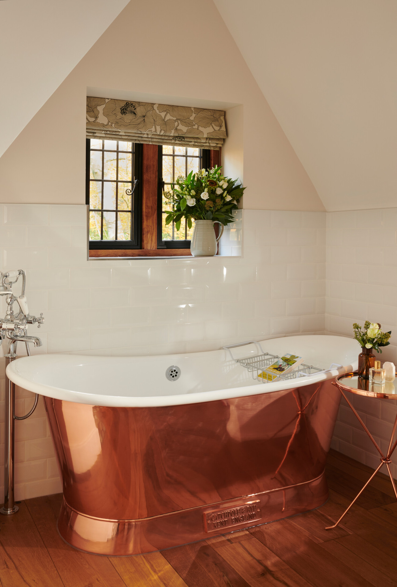 A cozy bathroom with a rose gold bathtub, window with a plant, and a small side table.