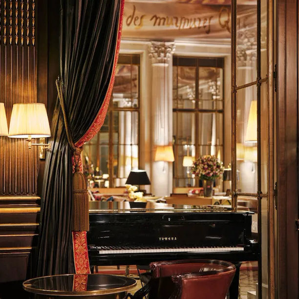 View from an English bar with wooden walls on a black piano. In the background you can see a restaurant with a brown toned canva on the ceiling. In the front of the picture you can see a round table with a leather chair. The piano is placed in a window door which is open. A heavy curtain with trimmings frames the window door, at Le Meurice, Paris