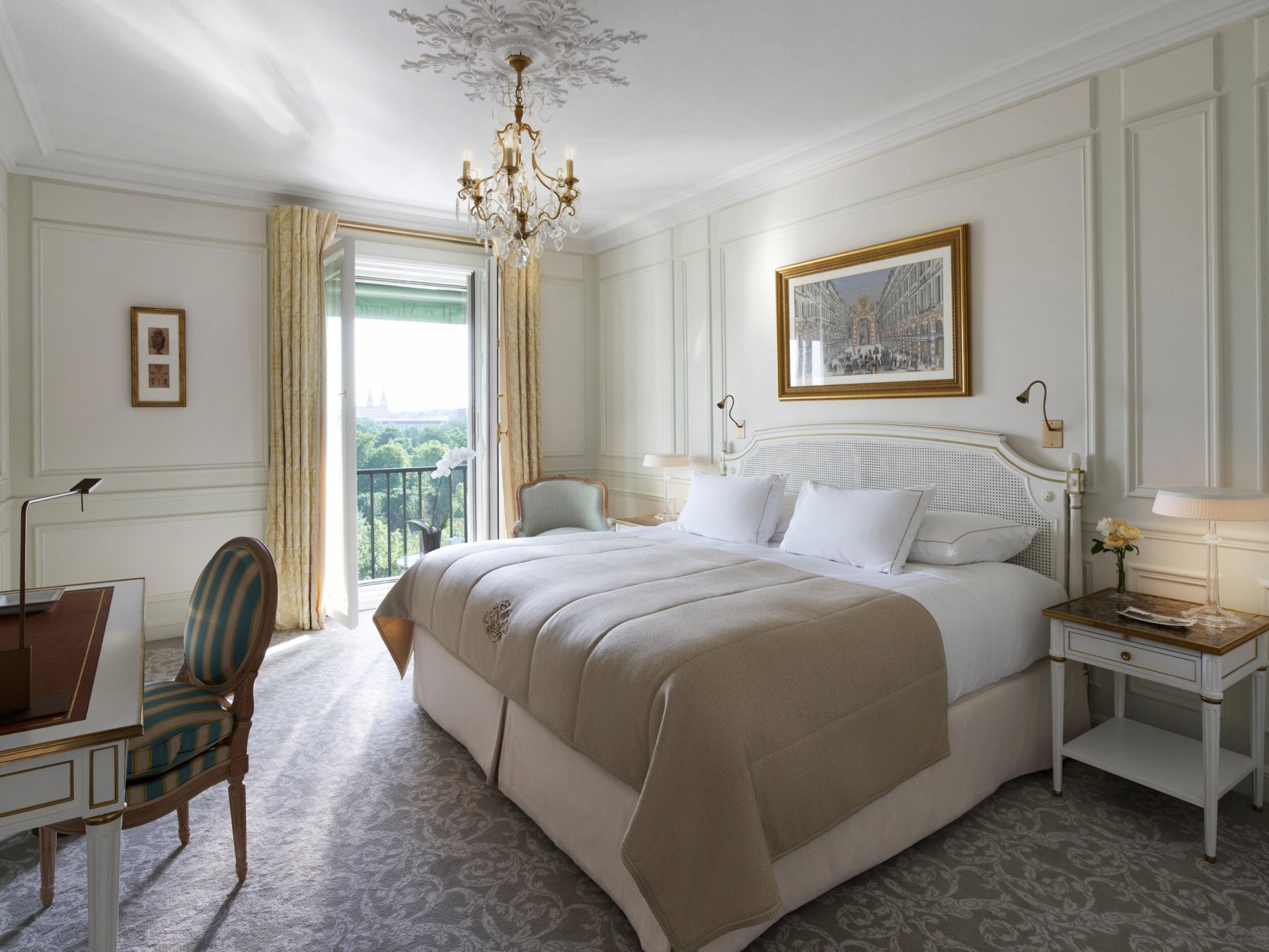 Luxurious bedroom with a large bed, beige bedding, and white pillows. A chandelier hangs from the ceiling. Open balcony doors reveal a lush view.
