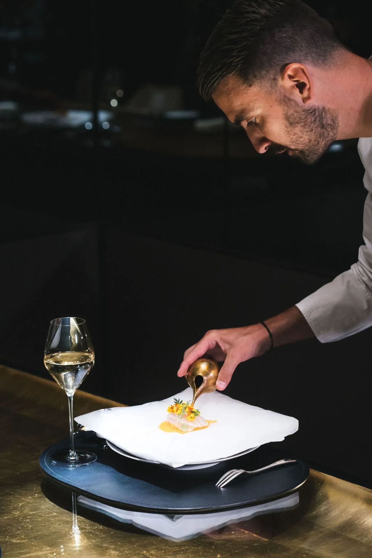 A chef is plating a delicate dish with precision using a brass tool. A glass of white wine sits on the table, creating an elegant and refined atmosphere.