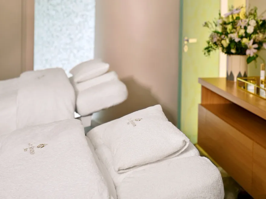 Spa room with a reclining massage chair covered in white towels. The soft glow of ceiling lights creates a serene ambiance. A bouquet of flowers sits on a wooden counter, adding elegance.