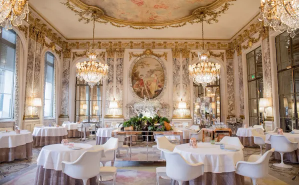 Luxurious dining room with ornate chandeliers, elegant round tables, and white chairs. Large murals and decorative gold accents create an opulent ambiance.