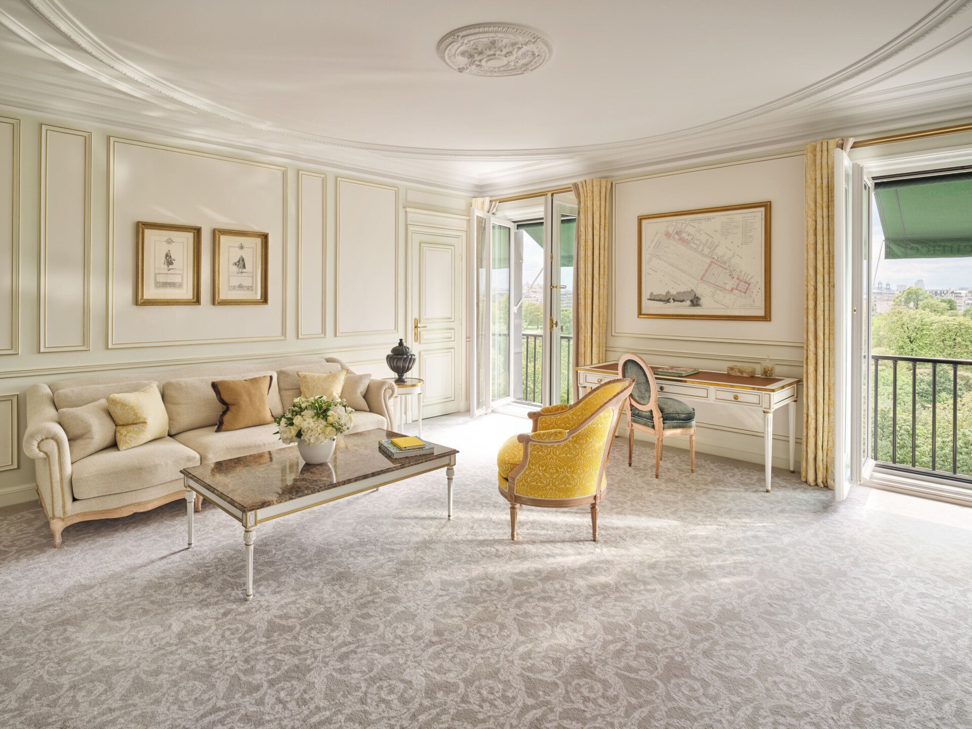 Living room with a beige canpé and a yellow sofa. Two big windows show the view on the Tuilerie garden, a office table and a grey blue chair is placed newt to the windows, at Le Meurice, Paris