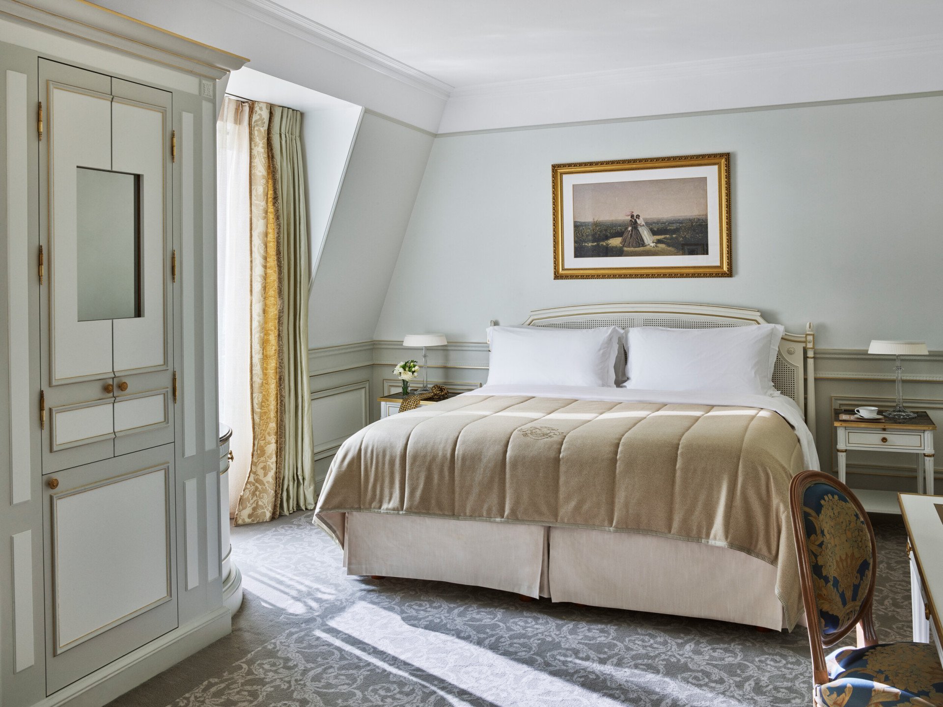 Luxurious hotel room featuring a neatly made king-sized bed with beige linens, elegant light blue walls, a framed artwork above, and soft lighting.