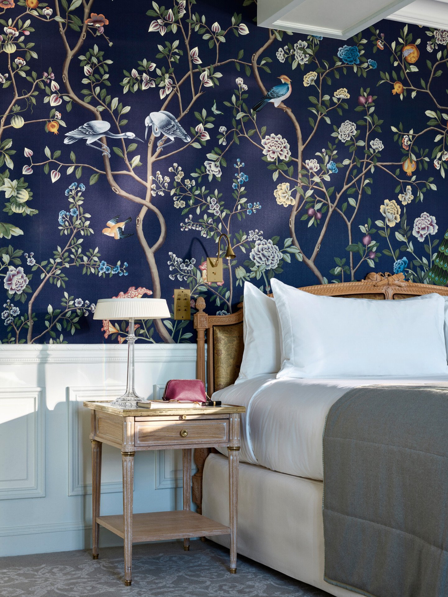 Portrait shot of the bedside table in an executive room, with a pouch and a book placed on the table. The bed highlights the dark blue hand painted wall. At Le Meurice, Paris.