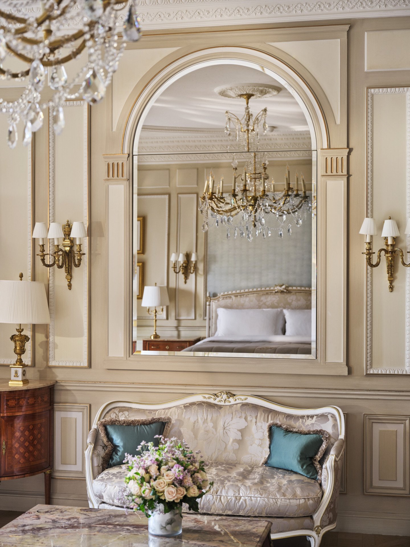 View on the table made with stone marquetry and a couch with a mirror above, which reflects the chandelier and the bed, at Le Meurice, Paris