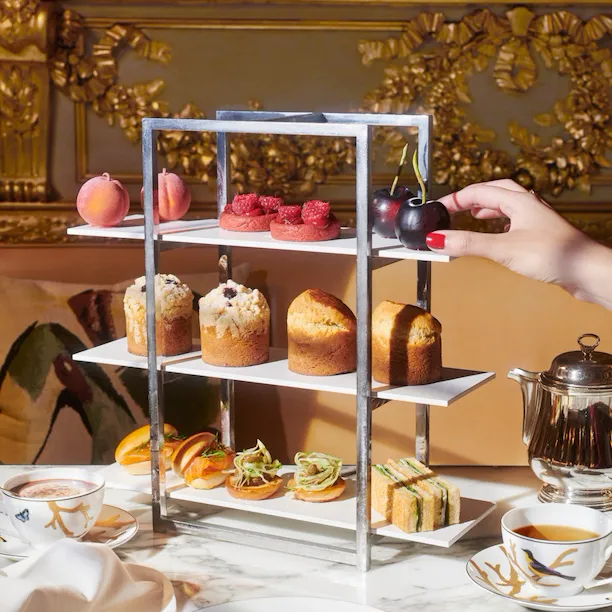 In the foreground, a plate with cultery and two cups of coffee and in the center, a display of cakes with brioche, snaks and fruit by Cédric Grolet that brings a lot of color to the photo, at Le Meurice, Paris