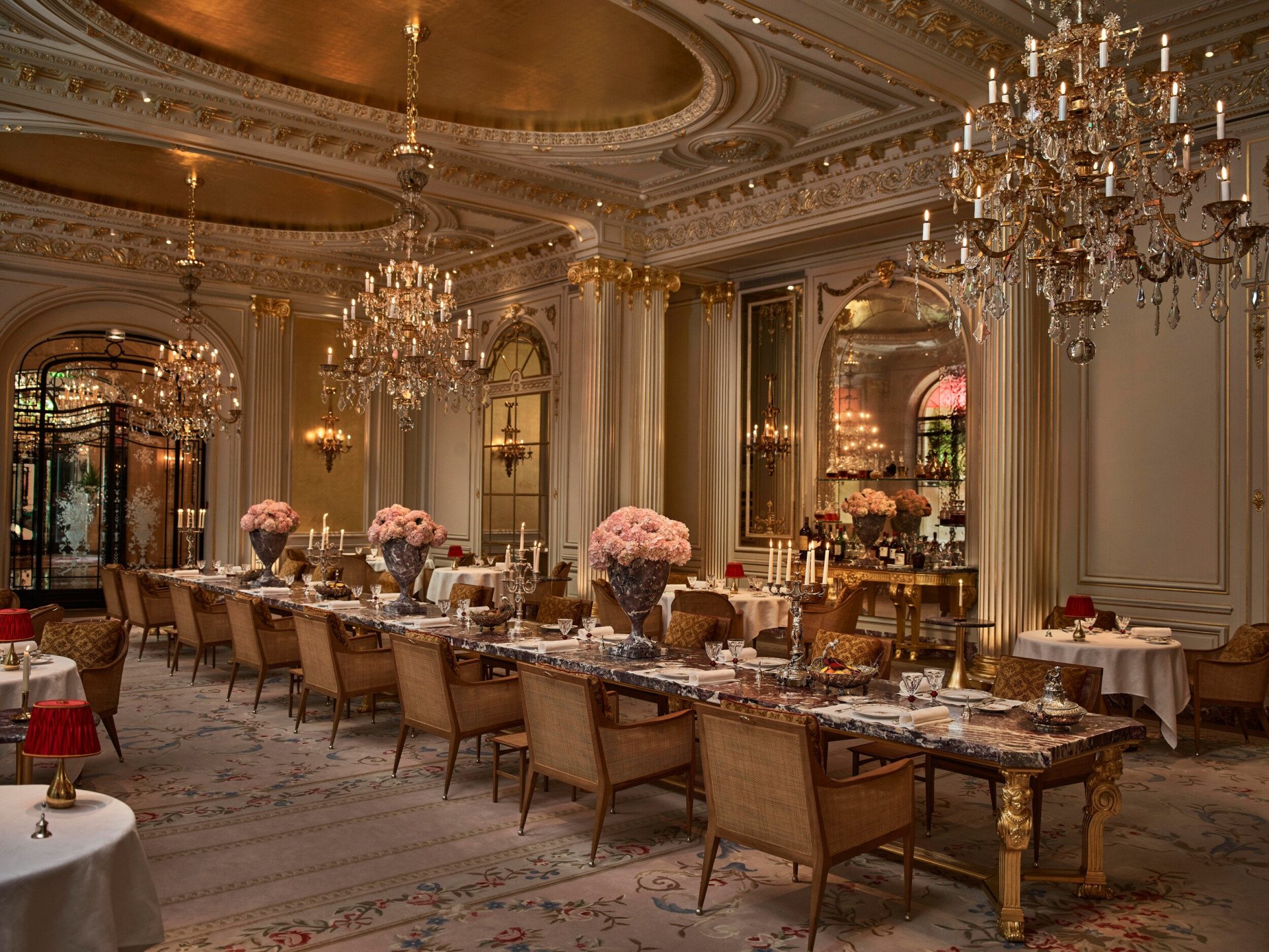 Overview of the Jean Imbert au Plaza Athénée Dining Room and its central marble table with flowers and crystal chandeliers hanging from the ceiling. At Hotel Plaza Athénée, Paris
