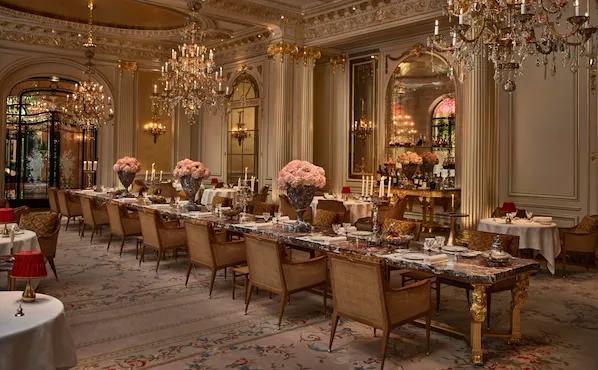 Overview of the Jean Imbert au Plaza Athénée Dining Room and its central marble table with flowers and crystal chandeliers hanging from the ceiling. At Hotel Plaza Athénée, Paris
