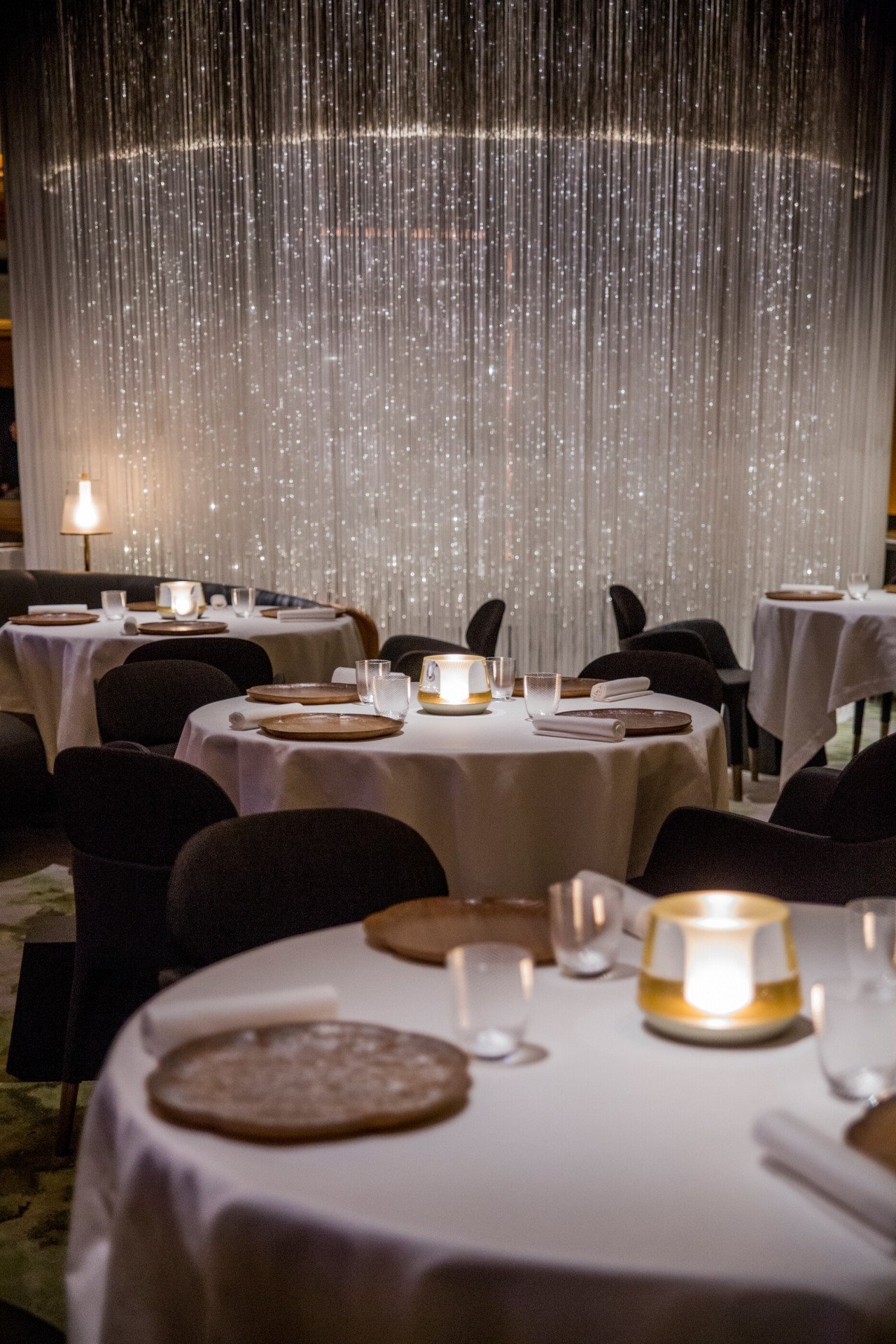 Alain Ducasse at The Dorchester Table details night time with dim lighting, various tables and table lumiere in background