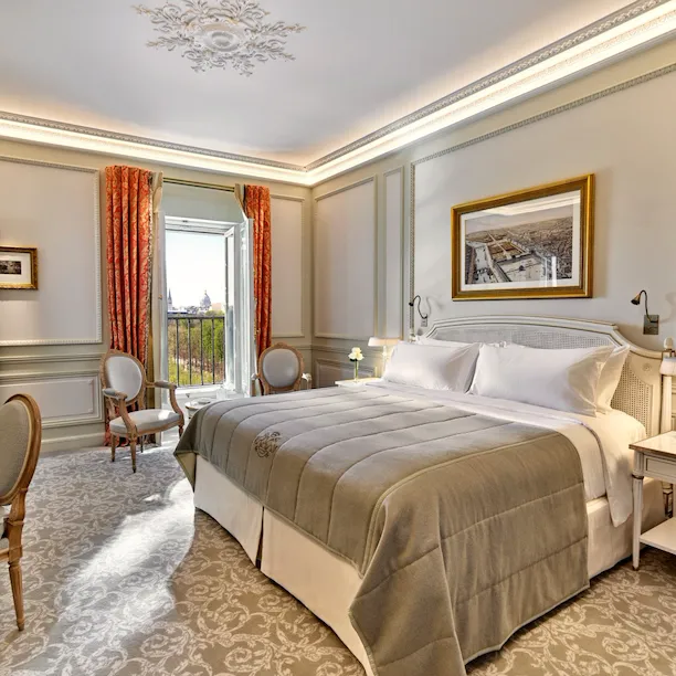 Luxurious hotel room with a plush bed, elegant chairs, and an open window with a scenic view. The room has ornate decor and warm, inviting tones.