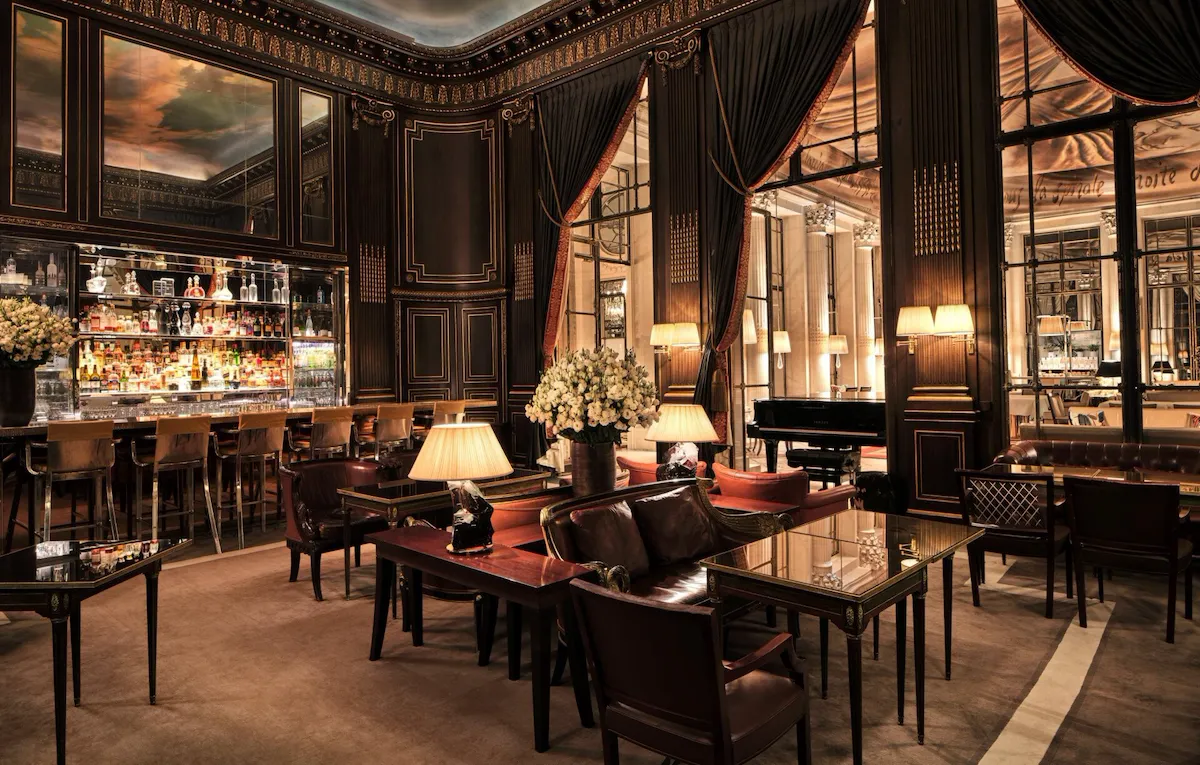 In the center of the photo, we see lamps lit on tables with armchairs and dark red cushions, and a bouquet of white flowers in a vase. On the left is bar 228 with high chairs. We can see the bottles on the shelves. Above the bar is a mirror reflecting the ceiling, which represents a sky with clouds. We look at the woodwork on the walls, with its gilded details. To the right is a piano and large windows with red curtains, revealing another room at Le Meurice, Paris.